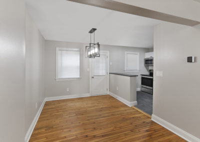 Living room facing the open kitchen with a lighting chandelier over the living room near the main entrance and window on hardwood-style flooring