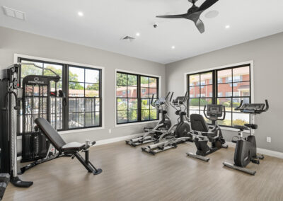 Fitness center at Jean Maria Gardens with large windows for natural lighting and both muscle and cardio equipment on hardwood-style flooring.